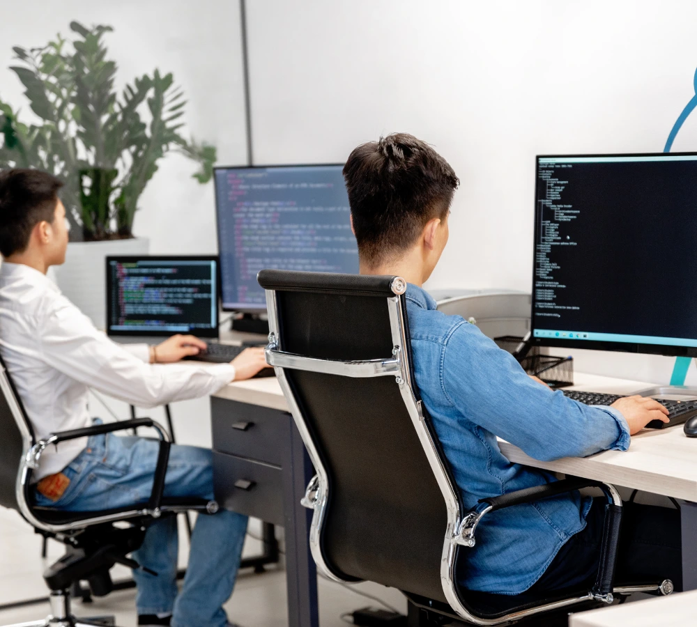 Valencia interior 2 Un programador sentado en un escritorio de oficina, de espaldas a la cámara, concentrado en un monitor grande que muestra código técnico, representando el desarrollo y la implementación continua de soluciones de asistente ia para soporte tic.